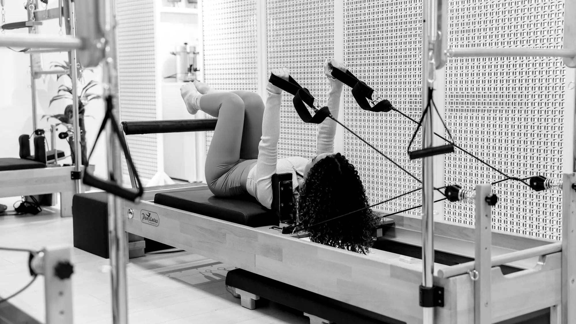 Woman using leg straps on a Pilates reformer beside a tower frame